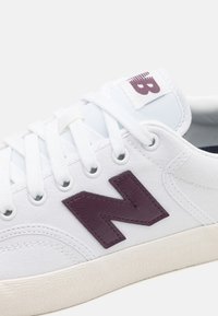 Baskets blanches avec logo "N" violet foncé, lacets blancs et semelle couleur crème, vue en gros plan sur le côté et la zone supérieure des lacets.