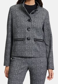 Giacca a quadri grigia con colletto alto, quattro bottoni neri e tasche con zip. Tessuto texture con una forma strutturata.