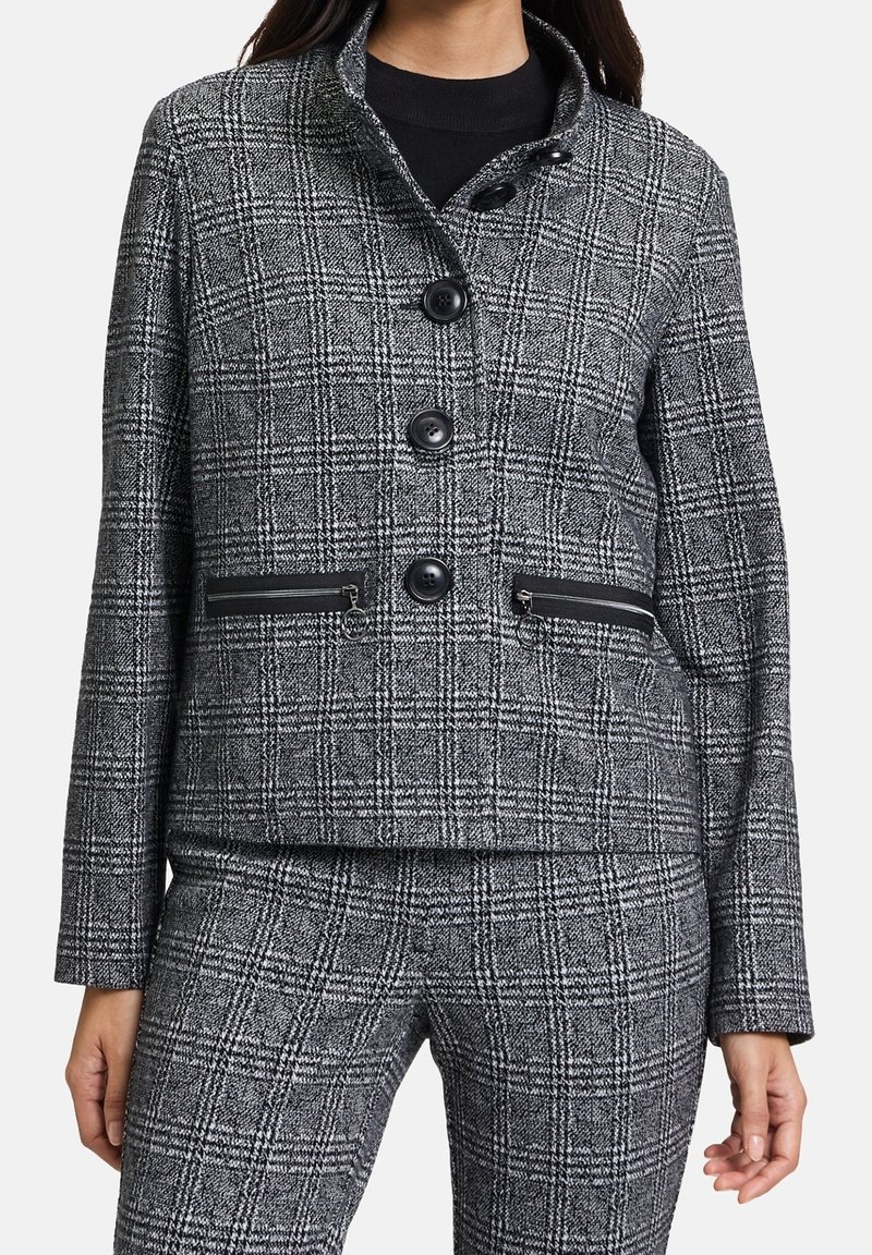 Giacca a quadri grigia con colletto alto, quattro bottoni neri e tasche con zip. Tessuto texture con una forma strutturata.