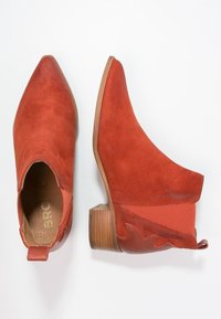 Botas de tobillo de ante rojo con punta afilada, un tacón de madera en forma de bloque y paneles laterales decorativos que presentan un sutil patrón ondulado.