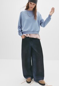 Felpa oversized azzurro chiaro, sovrapposta a un top rosa pastel, abbinata a pantaloni a gamba larga in denim scuro e scarpe stringate marroni.
