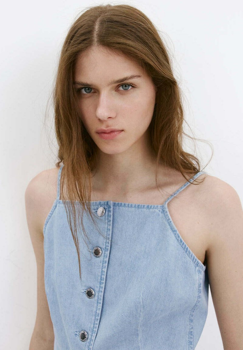 Jeune femme aux longs cheveux bruns portant un haut en denim bleu clair sans manches avec des boutons, face à un fond blanc uni.