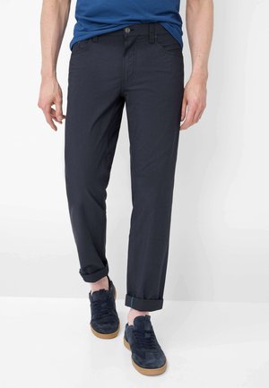 Mand iført opkørte mørkeblå bukser, navy sneakers med gummisåler og en blå t-shirt stoppet ned i bukserne, stående mod en hvid baggrund.