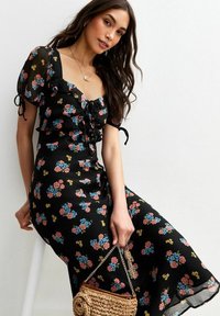 Robe noire à fleurs avec des manches courtes bouffantes, motif floral en rose et bleu, taille cintrée avec un détail à lacets, tissu texturé, jupe fluide.