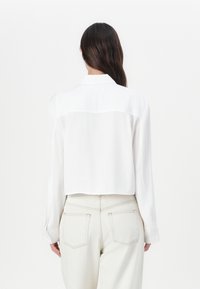 White collared shirt with long sleeves, cropped design, and smooth texture. Paired with light-colored jeans featuring double back pockets.
