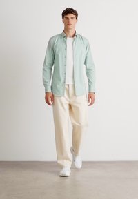 Cămașă butonată de culoare verde deschis peste un tricou alb, asociată cu pantaloni de culoare crem și adidași albi. Material neted, guler standard, fit lejer.
