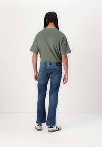 Vihreä lyhythihainen t-paita, vaaleaa kulutusta sinisissä farkuissa, laajeneva lahje ja mustavalkoiset raidalliset sandaalit, nähtynä takaa.