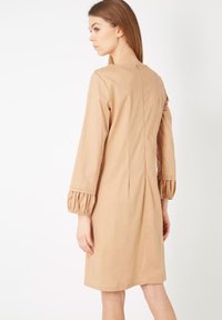 Vestido de algodón beige con un corte holgado, mangas largas con puños fruncidos y detalle de costura en la parte posterior. Textura suave.