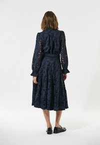 Navyblaues Spitzenkleid mit langen Ärmeln, floralem Muster, tailliertem Bund und fließendem Rock. Getragen mit schwarzen Slip-on-Schuhen.