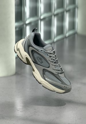Gray and white sneaker with mesh and leather panels, floating above a polished concrete floor near glass block wall background.