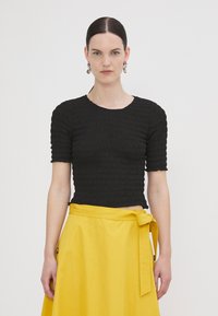 Top nero a maniche corte arricciato con scollo rotondo, abbinato a una gonna gialla brillante a vita alta con cintura annodata e dettagli di bottone.