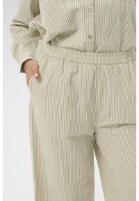 Personne portant un pantalon à rayures verticales beige et blanc et une chemise assortie, la main placée négligemment dans la poche.