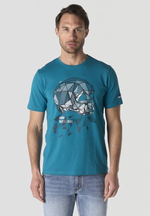 Hombre vestido con camiseta de color azul verdoso con diseño geométrico de calavera y texto, combinado con vaqueros azul claro, de pie sobre un fondo claro y liso.