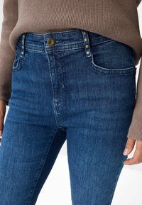 Jean en denim bleu taille haute avec une texture légèrement décolorée. Doté d'une fermeture à bouton et de clous décoratifs en métal sur la taille.