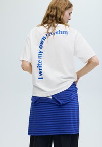 Weißes Baumwoll-T-Shirt mit blauem Schriftzug "I write my own rhythm" auf der Rückseite. Kombiniert mit einem blau-navy gestreiften Rock.