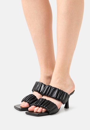 Mules pretos de salto alto com ponta quadrada e duas alças largas e franzidas feitas de material suave, texturizado para um detalhe adicional.