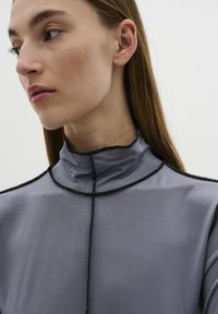 Graues hochgeschlossenes Oberteil mit transparenter Textur, das über schwarze Nähte am Ausschnitt und den Schultern verfügt und so das minimalistische Design betont.