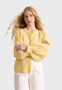 Blusa gialla a righe con scollo rotondo e maniche lunghe a sbuffo. Realizzata in tessuto leggero, presenta bottoni lungo la parte anteriore.