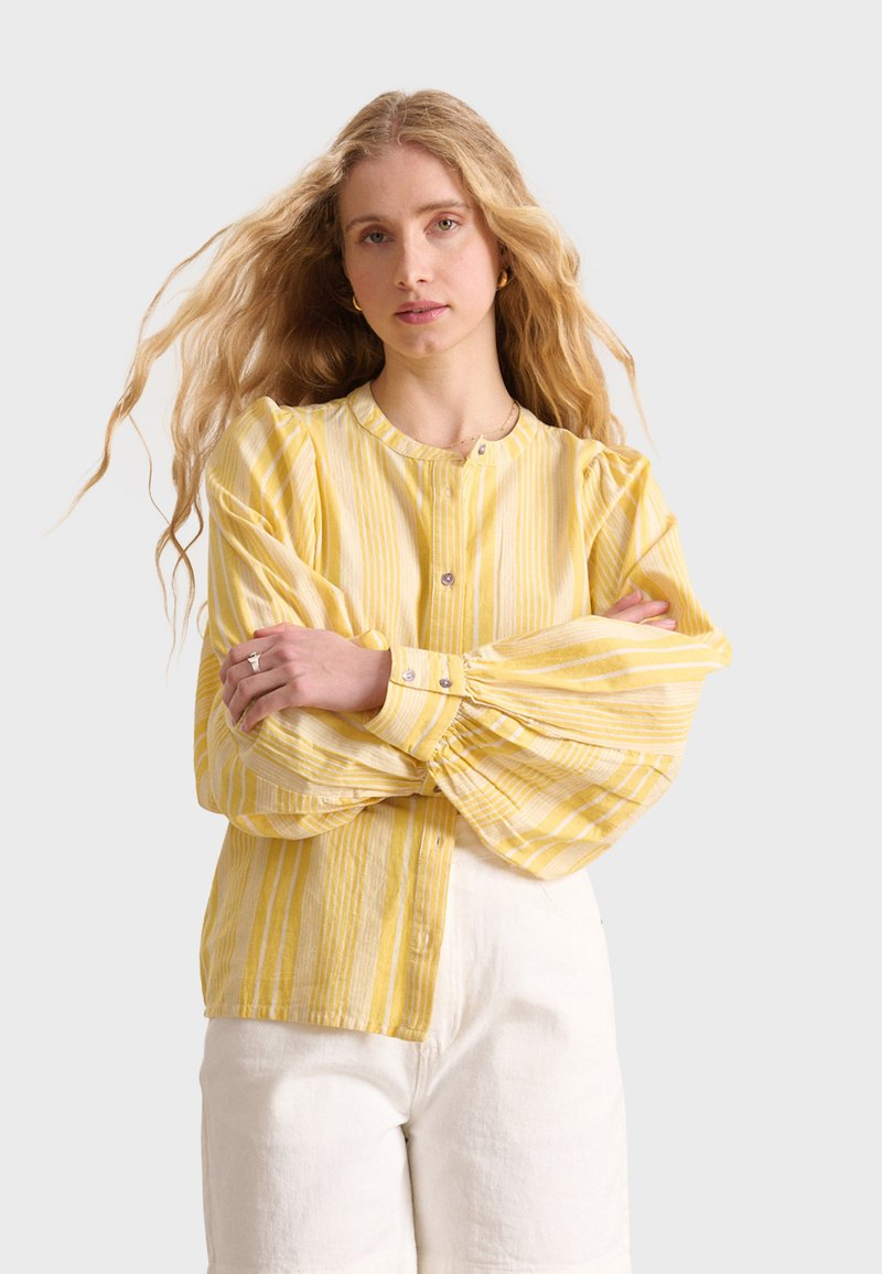 Blusa gialla a righe con scollo rotondo e maniche lunghe a sbuffo. Realizzata in tessuto leggero, presenta bottoni lungo la parte anteriore.