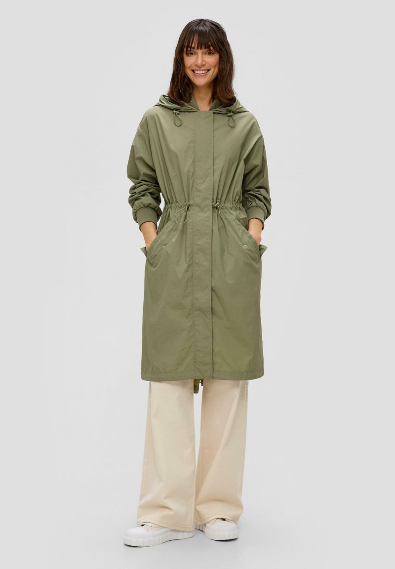 Olivegrüne, leichte Jacke mit Kapuze, Kordelzug in der Taille, langen Ärmeln und zwei Fronttaschen. Wird über cremefarbenen, weiten Hosen getragen.