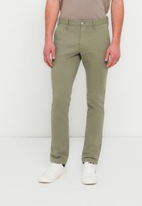 Χακί παντελόνια chinos από βαμβακερό μείγμα, με στενή γραμμή, μπροστινές τσέπες, κλείσιμο με κουμπιά και λείο φινίρισμα. Συνδυασμένα με λευκά αθλητικά παπούτσια.