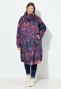 Impermeabile blu con motivi astratti rosa e rossi, dotato di cappuccio, tasca frontale e vestibilità ampia, abbinato a jeans scuri e stivali crema.