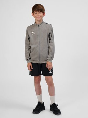 Dreng iført en grå sportstrøje med lynlås, sorte shorts, hvide sokker og sorte sneakers, stående og smilende mod en ensfarvet baggrund.