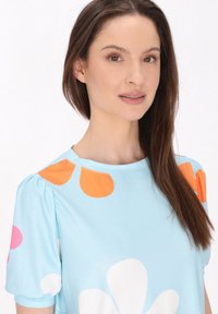T-shirt azzurro chiaro con grandi motivi floreali colorati in rosa, bianco e arancione. Maniche a sbuffo corte, tessuto morbido, scollo rotondo.