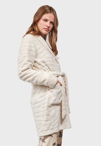 Witte pluche badjas met gestructureerde strepen, met een riem in de taille. Voorzien van een sjawlkragen en zachte, donzige manchetten. Twee voorzakken zichtbaar.