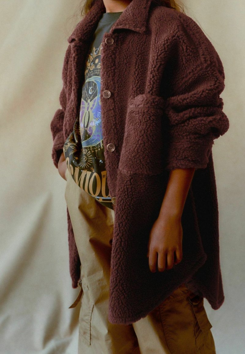 Personne portant un manteau marron duveteux sur un t-shirt graphique et un pantalon beige, debout devant un fond neutre uni.