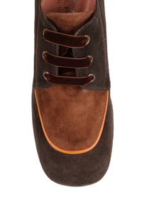Brązowe zamszowe buty z ciemniejszą brązową górą, jaśniejszym brązowym przodkiem, pomarańczowym akcentem na krawędzi oraz trzema brązowymi sznurówkami.