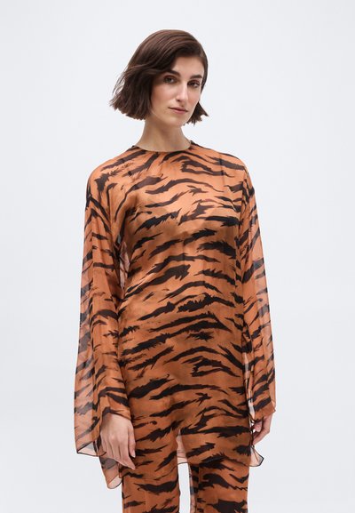 Γυναίκα που φοράει διάφανο πορτοκαλί τοπ με μακριά μανίκια, διακοσμημένο με μαύρες ρίγες τιγρέ, και παντελόνι που ταιριάζει, στέκεται μπροστά σε ένα απλό λευκό φόντο.