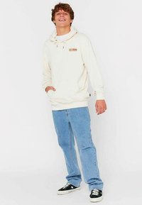 Cremefarbener Kapuzenpullover mit Fronttasche, kombiniert mit hellblauen Jeans und schwarzen Sneakers mit weißen Akzenten.