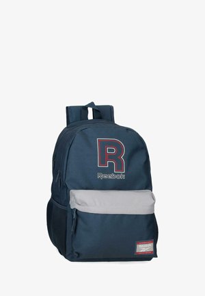 Mochila azul marino de Reebok con gran logo "R" en rojo y blanco, bolsillo frontal gris con cremallera, bolsillos laterales de malla y correas de hombro acolchadas.