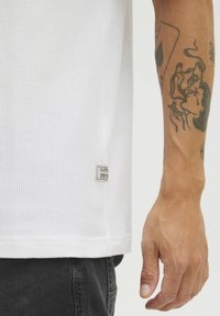 Hvid tekstureret skjorte med et lille logo-tag på den nederste søm; hånden, der bærer skjorten, har detaljerede tatoveringer.