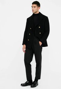 Črni dvoplastni volneni plašč z zlatimi gumbi, širokim ovratnikom, kombiniran s prilagojenimi črnimi hlačami in poliširanimi čevlji.