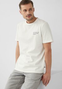 Korte mouw crème t-shirt van katoen, met een kleine afgedrukte tekst nabij de borst. Gecombineerd met lichtgrijze jeans.