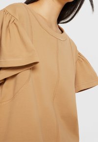 Haut court à manches courtes de couleur taupe avec des manches bouffantes lâches et texturées ainsi que des coutures contrastantes. Le tissu semble doux et ajusté autour de l'encolure.