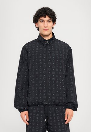 Black zip-up jacket featuring a subtle all-over pattern, elastic cuffs, and a high collar. The material appears lightweight and textured.