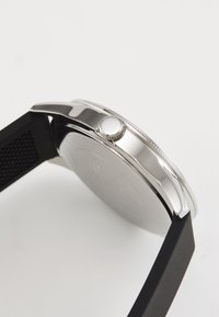 Reloj de metal plateado con bisel texturizado, caja redonda y correa de silicona negra. Incluye una corona de presión en el lateral.