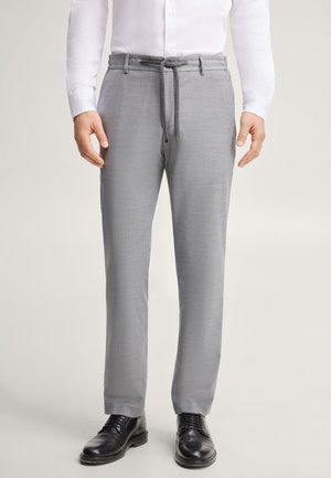 Uomo che indossa pantaloni grigio chiaro con coulisse in vita, camicia bianca abbottonata e scarpe eleganti nere, in piedi su sfondo semplice.