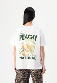 Valge puuvillane T-särk, mille seljal on suur virsiku graafika, mille all on roheline tekst "Just PEACHY" ja "Ja 100% LOODUSLIK."