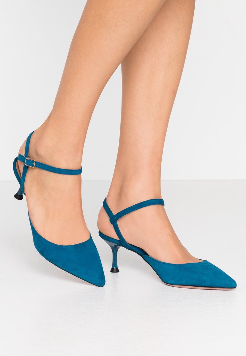 Pieds portant des escarpins slingback en cuir suédé bleu sarcelle à bout pointu, avec de fines brides à la cheville et des petits talons sur un sol blanc uni.