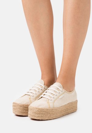 Beige platform sneakers med flettede jutesåler båret på bare fødder mod en ensfarvet lys baggrund.