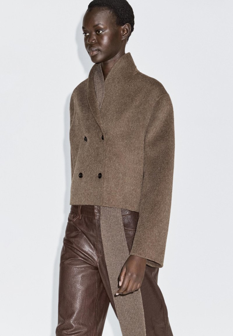 Brown wool blend cropped jacket with a double-breasted design, black buttons, and a shawl collar, paired with brown leather pants.