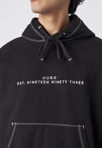 Homme portant un sweat à capuche noir avec des cordons blancs et le texte "HUGO EST. NINETEEN NINETY THREE" brodé sur la poitrine.