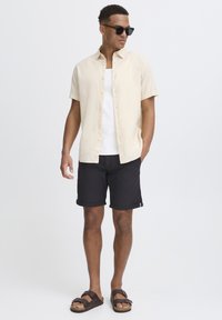 Camisa de manga corta abotonada en un color beige claro, combinada con pantalones cortos negros enrollados. Usada con una camiseta sin mangas blanca y sandalias marrones. Gafas de sol negras presentes.