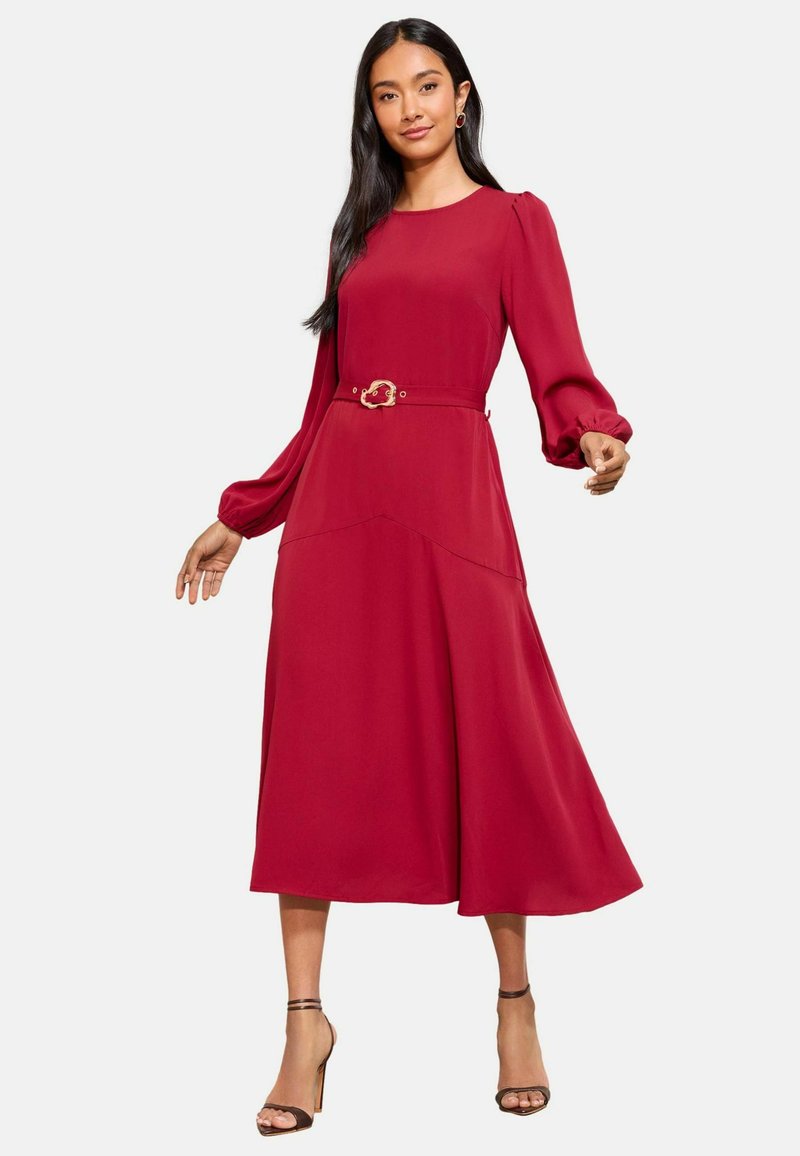 Rochie roșie cu mâneci lungi, cu un corset ajustat și fustă evazată. Prezintă un detaliu cu cataramă din aur și o țesătură moale și fluidă.