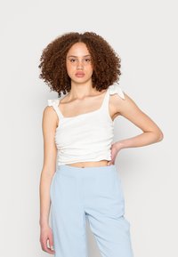 Top branco, curto, sem mangas, com detalhe torcido e laços nas alças do ombro; combinado com calças largas de tom azul claro.