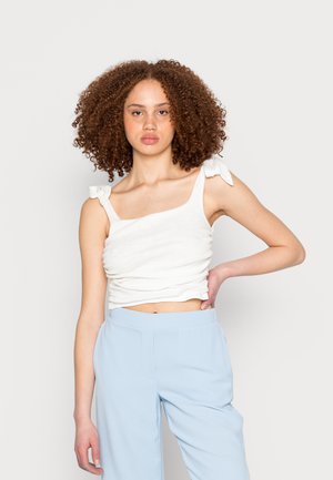 Top branco, curto, sem mangas, com detalhe torcido e laços nas alças do ombro; combinado com calças largas de tom azul claro.
